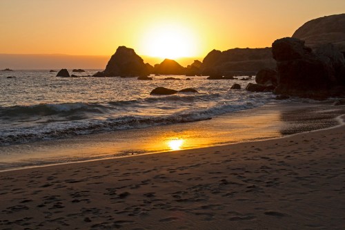 Sunset over a rocky beach, with golden light reflecting on the water and footprints in the sand.