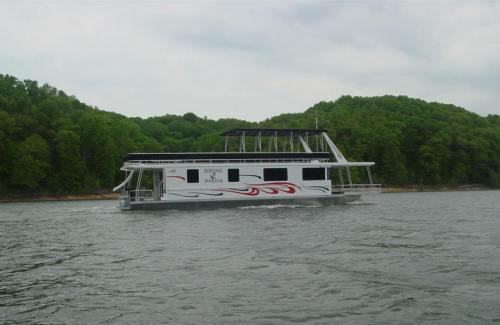 A houseboat with a sleek design glides across a calm lake, surrounded by lush green hills.