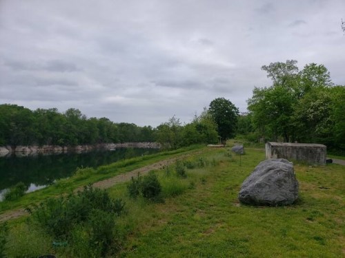 A serene landscape featuring a calm river, grassy banks, and large rocks under a cloudy sky.