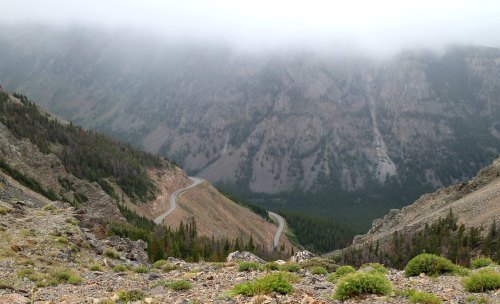 A winding road cuts through a mountainous landscape, shrouded in mist and surrounded by rugged terrain and greenery.