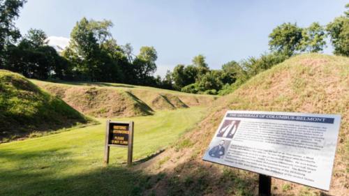 Historic site with grassy mounds and informational signs about the Generals of Columbus-Belmont. Trees in the background.