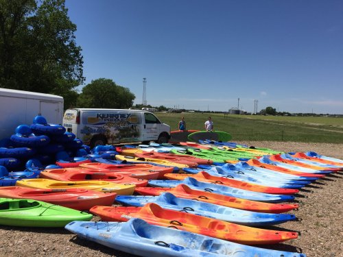 A variety of colorful kayaks lined up on the ground near a van, with a grassy field and blue sky in the background.