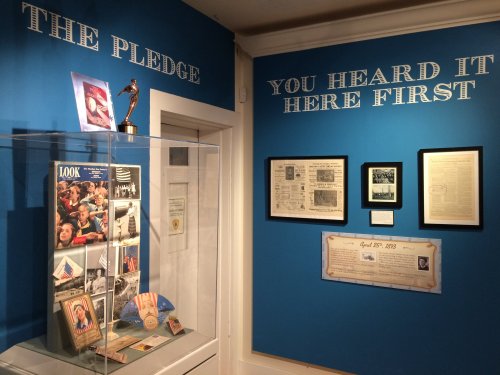 Exhibit featuring "The Pledge" with display cases, framed documents, and a blue wall with text.