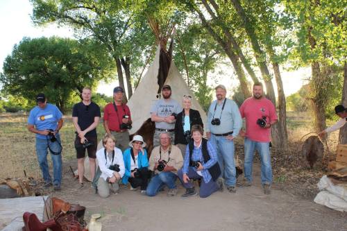 A group of photographers poses in front of a teepee, surrounded by trees and nature, with cameras in hand.