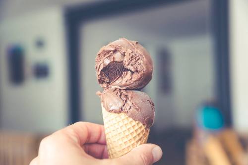 A hand holding a cone with two scoops of chocolate ice cream, one partially eaten, against a blurred background.
