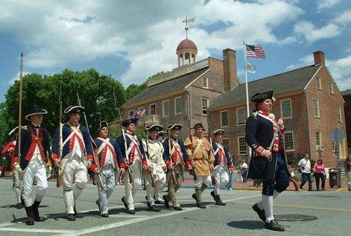 A group of historical reenactors in colonial uniforms march past a historic building on a sunny day.