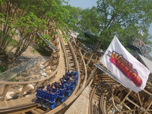 A wooden roller coaster named "Wooden Warrior" with riders on the track, surrounded by trees and a flag.