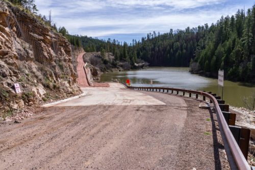 A dirt road leads to a calm lake surrounded by trees, with a red marker and signs visible along the roadside.
