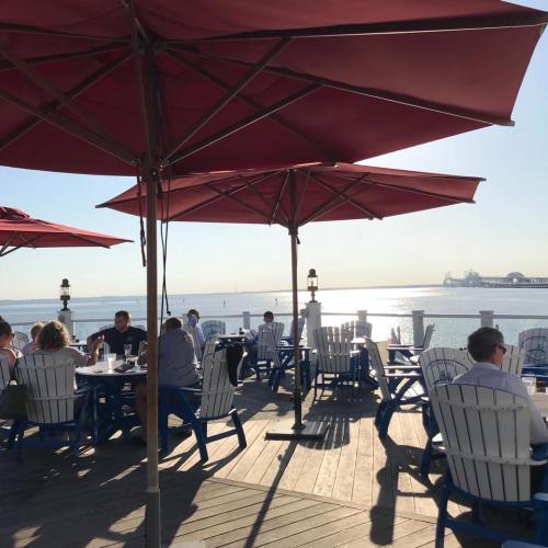 Outdoor dining area with red umbrellas, blue chairs, and people enjoying the view of the water and pier in the background.