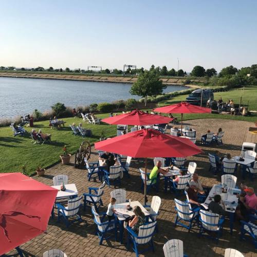 Outdoor dining area by a river, featuring blue chairs, red umbrellas, and people enjoying the sunny weather.