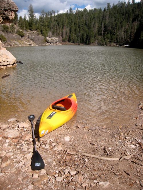 A bright yellow kayak rests on a rocky shore by a calm lake, surrounded by trees and a cloudy sky.