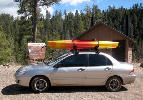 A silver car with a kayak on the roof parked near a sign for Coconino National Forest, surrounded by trees.