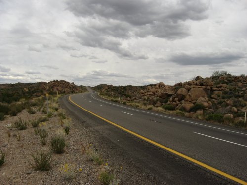 A winding road stretches through a rocky landscape under a cloudy sky. Sparse vegetation lines the roadside.