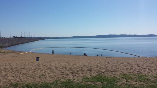 A sandy beach by a calm lake on a clear day, with a few people enjoying the water and distant hills in the background.