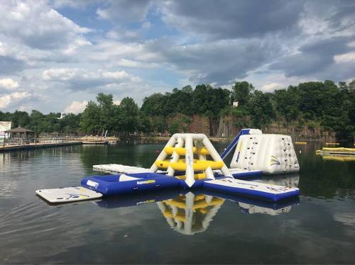 Inflatable water obstacle course on a lake, surrounded by trees and under a cloudy sky.
