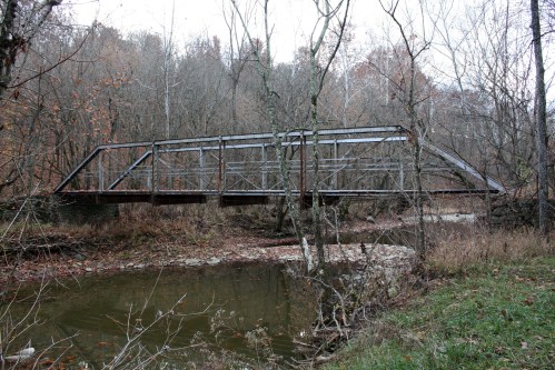 A metal truss bridge spans a small creek, surrounded by trees and autumn foliage.