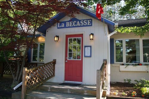 A charming restaurant entrance with a red door, blue trim, and a sign reading "La Bécasse" surrounded by greenery.