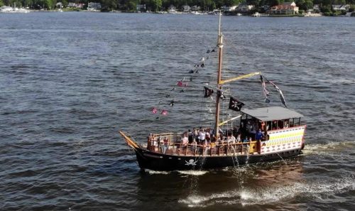 A pirate-themed ship sails on a river, with people on board and flags waving in the breeze.