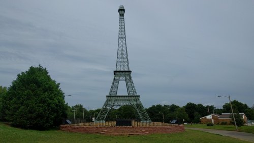 A replica of the Eiffel Tower stands in a park, surrounded by greenery and a cloudy sky.