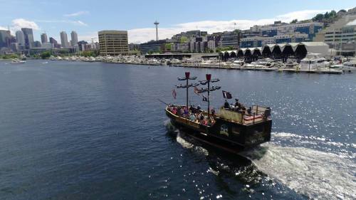 A pirate-themed boat sails through a harbor with city buildings and a space needle in the background.