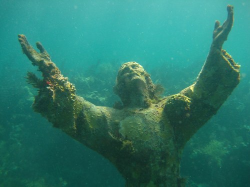 A submerged statue of Christ with outstretched arms, covered in marine life, surrounded by clear blue water.