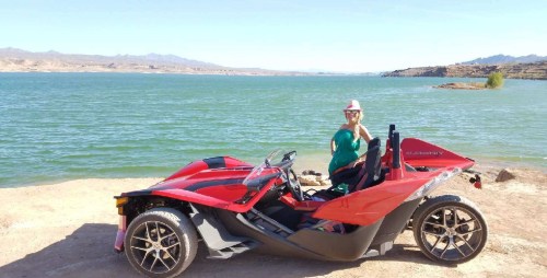 A person in a hat poses next to a red three-wheeled vehicle by a lake with mountains in the background.