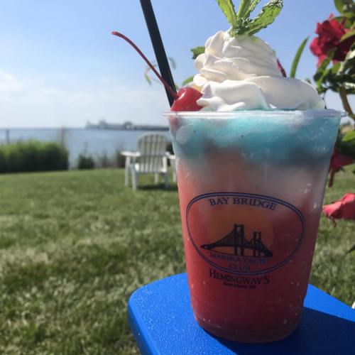 A colorful layered drink with whipped cream and a cherry, set against a scenic waterfront view.