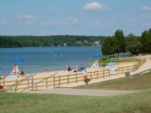 A sandy beach by a lake with umbrellas, lifeguard stands, and people enjoying the sun and water on a clear day.