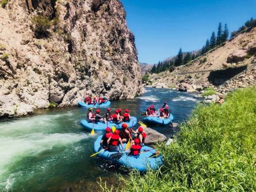 A group of people in red helmets rafting on a river surrounded by rocky cliffs and greenery under a clear blue sky.