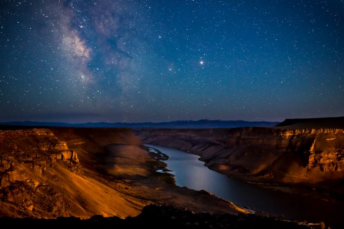 A serene night sky filled with stars above a winding river and rugged cliffs, illuminated by the Milky Way.