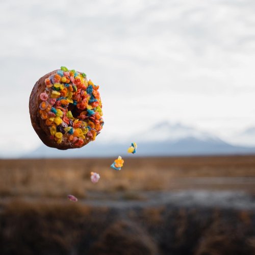 A colorful donut floats in the air, surrounded by scattered cereal pieces, with a blurred landscape in the background.