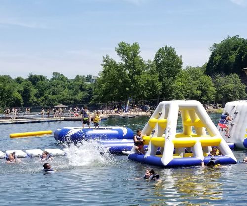 A sunny day at a lake with inflatable water structures and people swimming and playing. Trees and a dock in the background.
