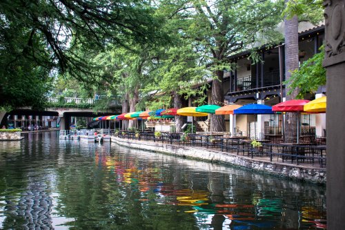 Colorful umbrellas line a riverside patio, reflecting in the calm water surrounded by lush greenery.