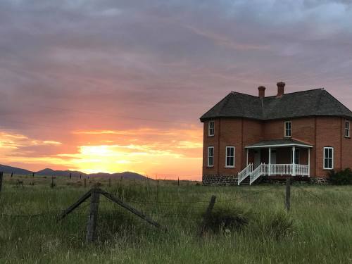 A historic brick house stands in a grassy field, framed by a vibrant sunset and distant mountains.