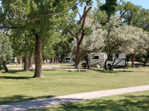 A grassy park with trees, featuring an RV parked in the background and a walking path in the foreground.