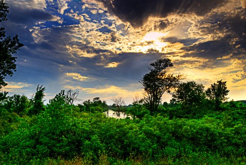 A vibrant landscape featuring lush greenery, a river, and a dramatic sky with clouds and sunlight breaking through.