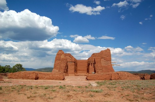 Ruins of a red adobe structure against a blue sky with clouds, surrounded by green grass and distant mountains.