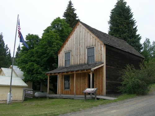 A rustic wooden building with a sloped roof, surrounded by trees, and an American flag nearby.