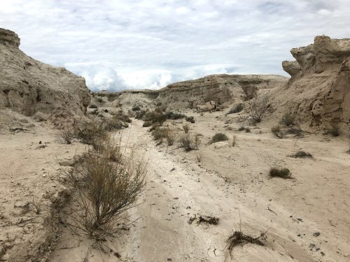 A barren landscape with dry, cracked earth, sparse vegetation, and rocky formations under a cloudy sky.