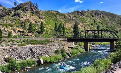A scenic river flows under a bridge, with a group rafting and lush green hills in the background under a blue sky.