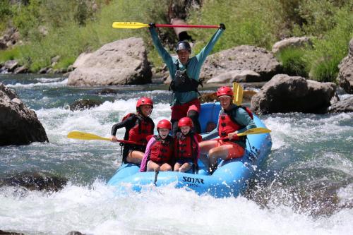 A group of five people in a blue raft, smiling and paddling through a river with splashing water.