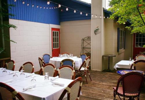 Outdoor dining area with white tables, wicker chairs, and string lights, surrounded by greenery and colorful building walls.