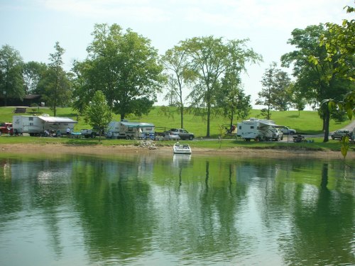 A serene lakeside scene with RVs parked along the shore and a boat on the water, surrounded by lush greenery.