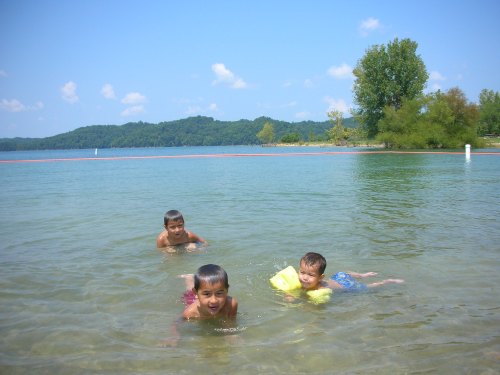 Three young boys play in shallow water at a lake, enjoying a sunny day with green hills in the background.