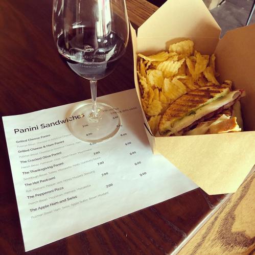 A menu with panini options, a glass of red wine, and a takeout box of a sandwich with chips on a wooden table.