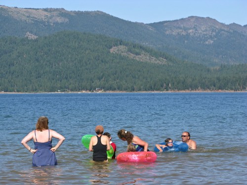 A group of people enjoying a sunny day at the lake, playing in the water with colorful floaties and mountains in the background.
