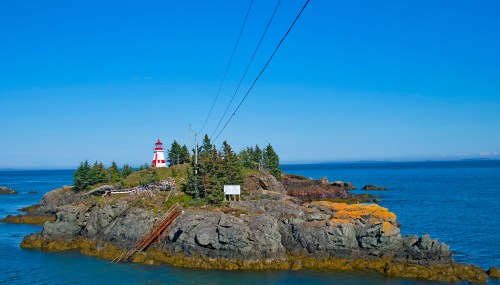 A red and white lighthouse on a rocky island surrounded by blue water and green trees under a clear blue sky.