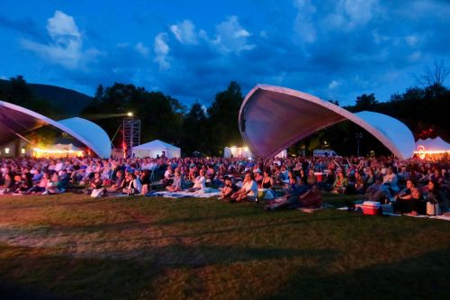 A large crowd sits on the grass at an outdoor concert, with tents and dramatic clouds in the evening sky.