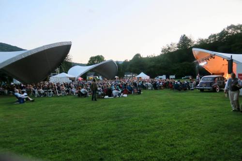 A large crowd gathers on a grassy field for an outdoor concert, with tents and a vintage car in the foreground.
