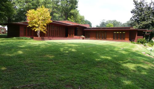 A modern wooden house with large windows, surrounded by green grass and trees on a sunny day.